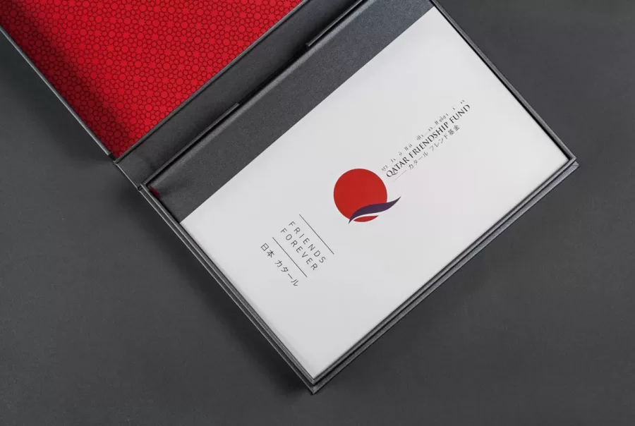 Open box with red patterned interior, showing a booklet with "Qatar Friendship Fund" and "Friends Forever" text, and a red circle logo.